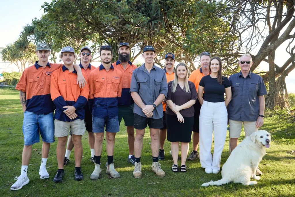 The Ocean Blue Metal Roofing team standing together on the grass under a pandanus tree by the beach, with the team dog