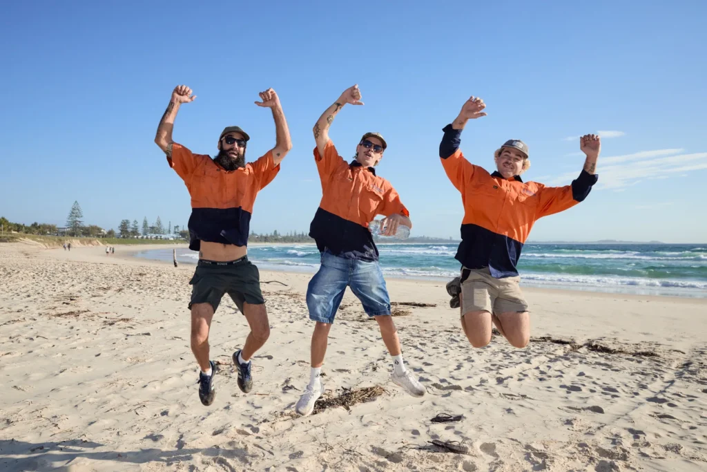 Three Ocean Blue Metal Roofing team members in orange and navy hi-vis shirts jumping in the air on the beach