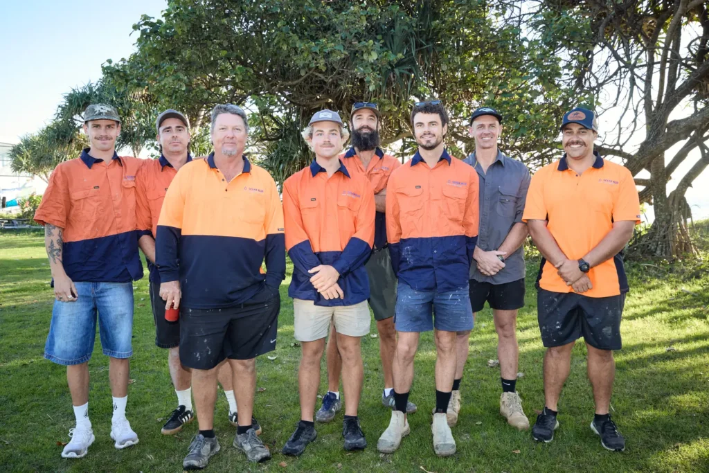 Ocean Blue Metal Roofing roofers in orange and navy hi-vis work shirts standing together outdoors under a pandanus tree